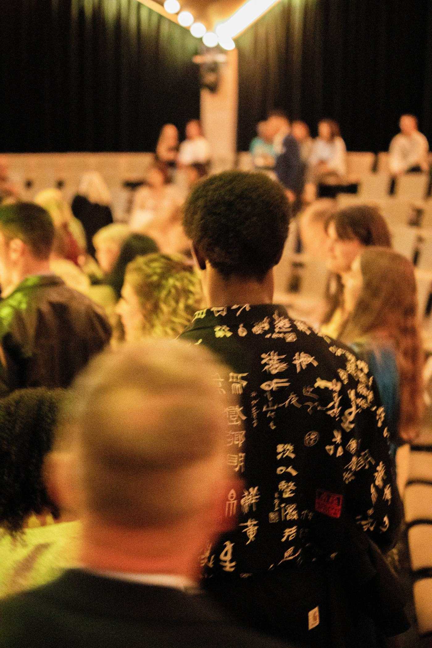 An audience seen from within the crowd, faces lit from the stage