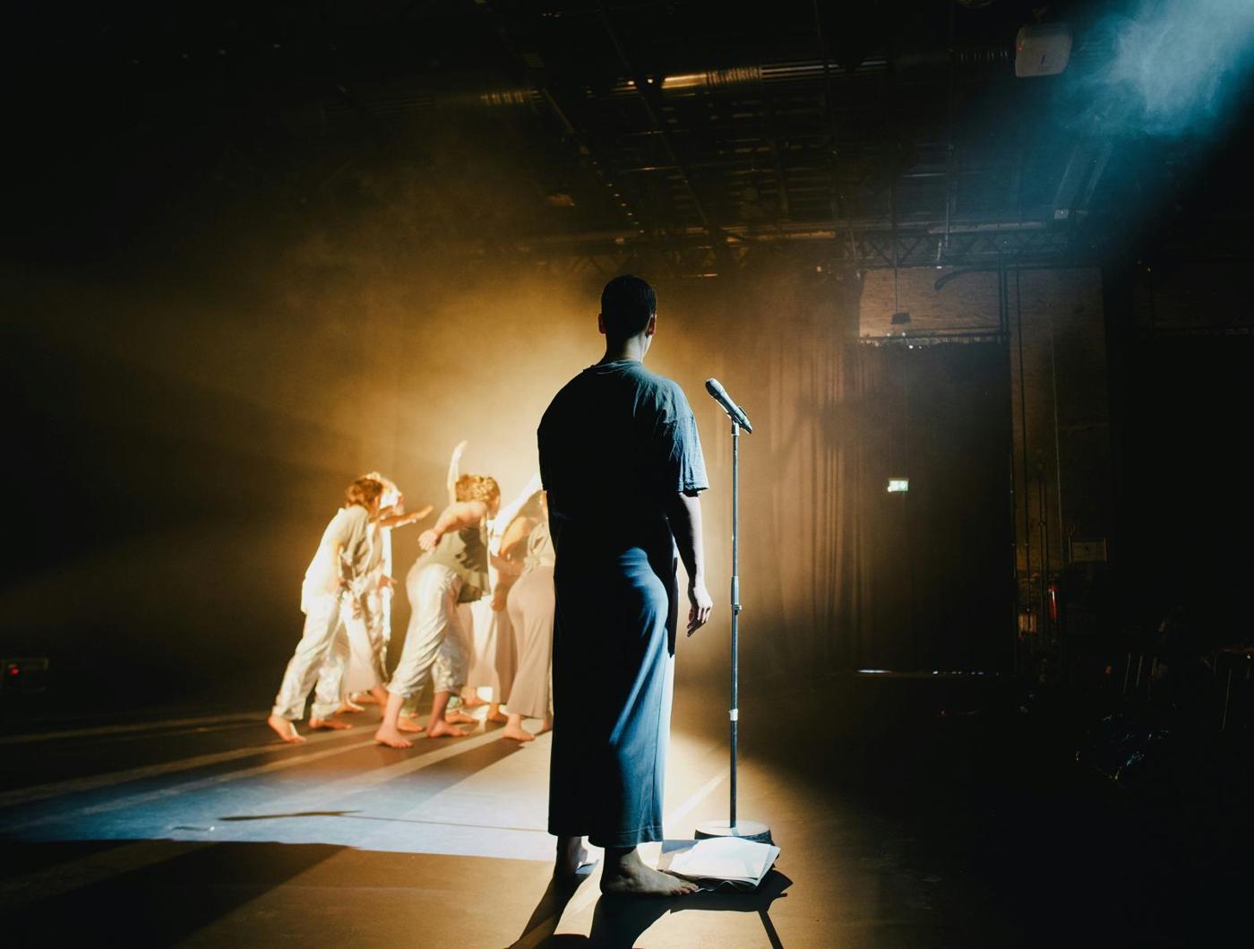 A performer standing at a microphone on a dark stage, with dancers in white behind them