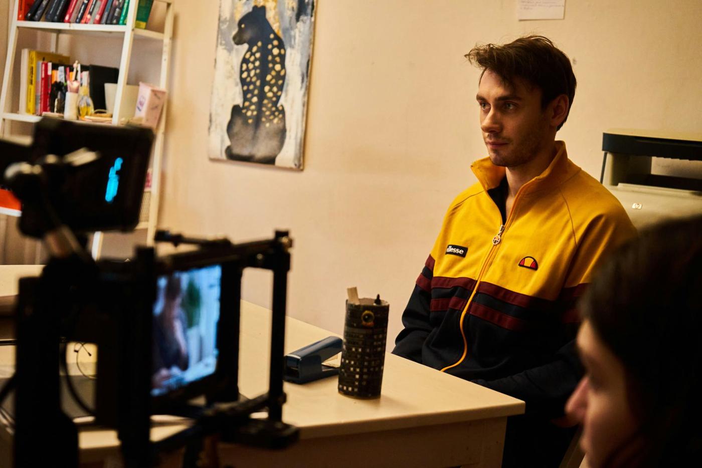 A person seated at a desk facing a camera on a tripod