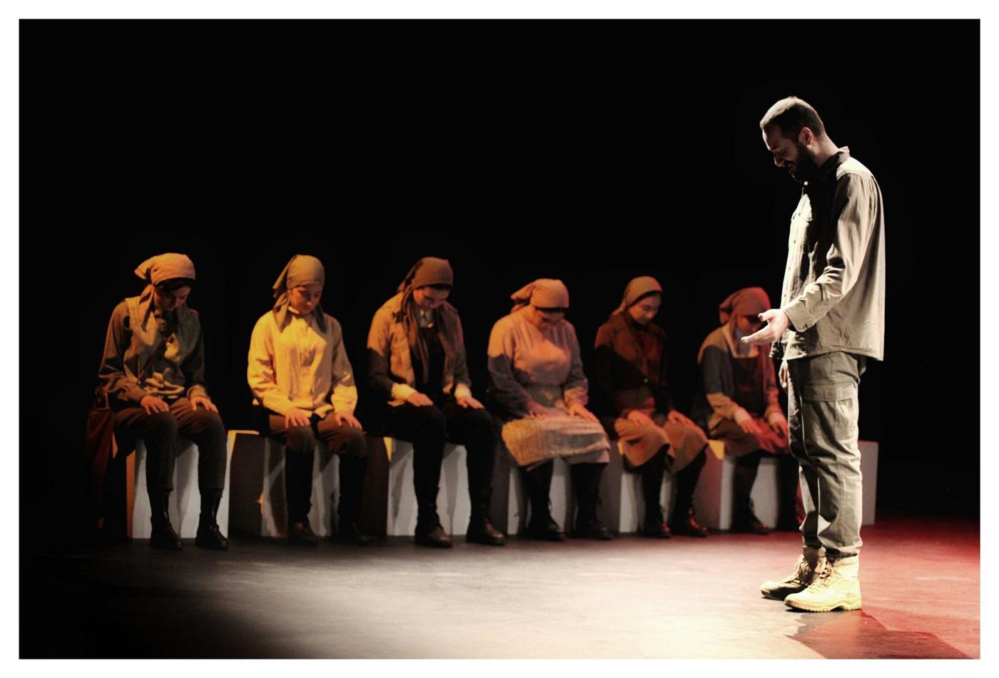 A row of people seated along the front of a stage, watching one person standing